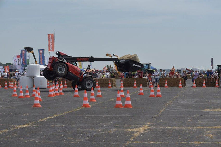 Machine demonstration with tractor at outdoor trade fair, surrounded by spectators, hay bales, and banners.