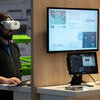 Person mit VR-Headset an einem Messestand mit Monitoren, Infografiken und Laptop.